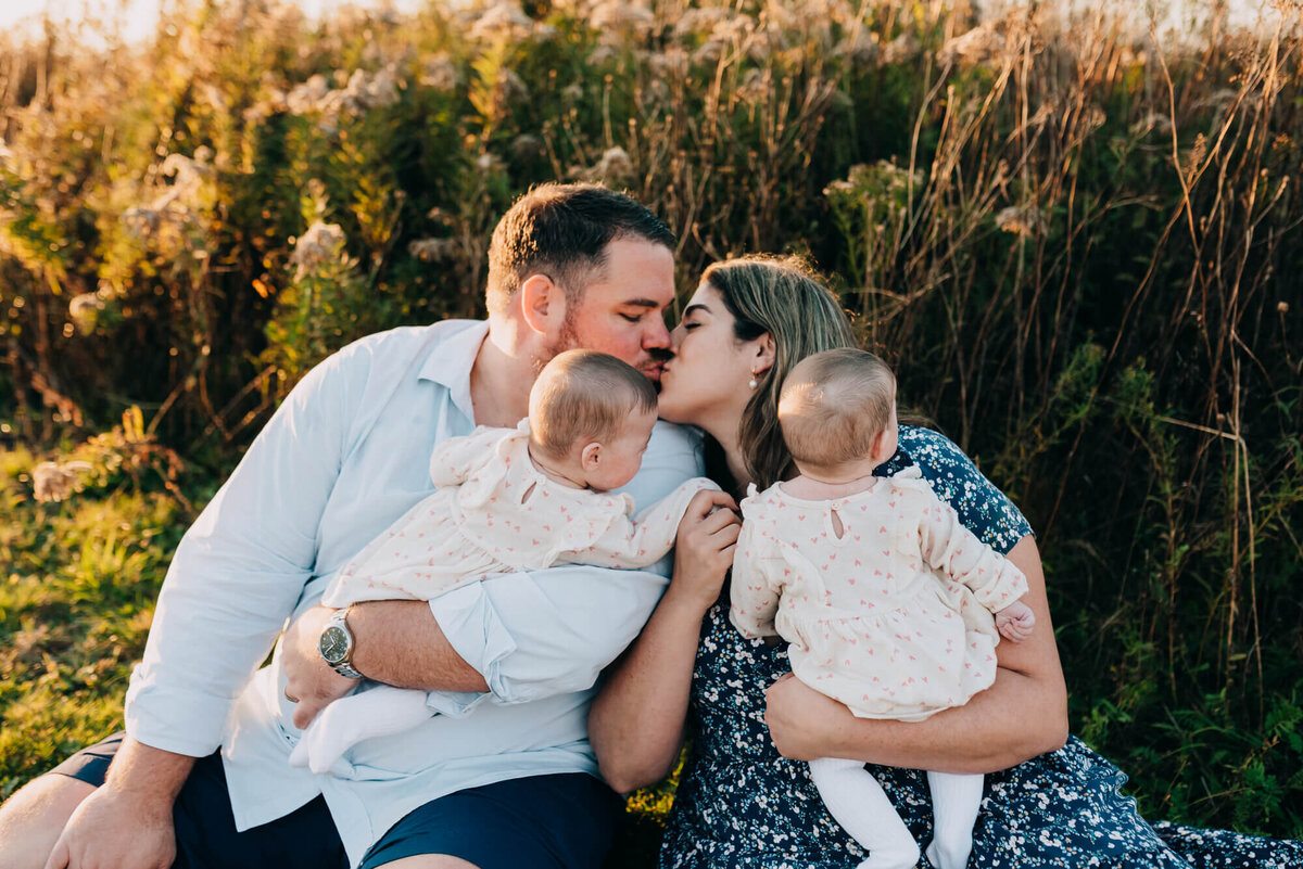 A mother and father hold their twin baby girls and give each other a kiss during sunset
