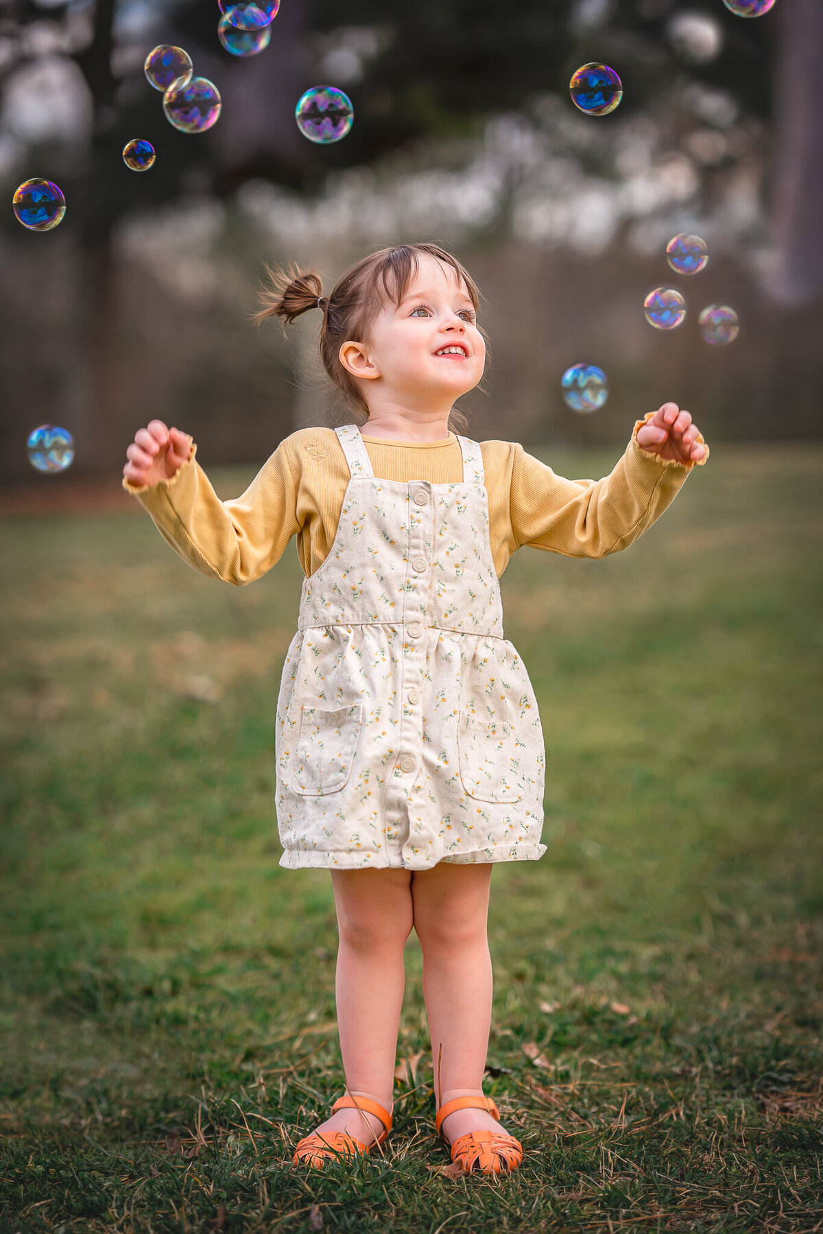 A little girl in a yellow shirt is looking at bubbles floating though the air.