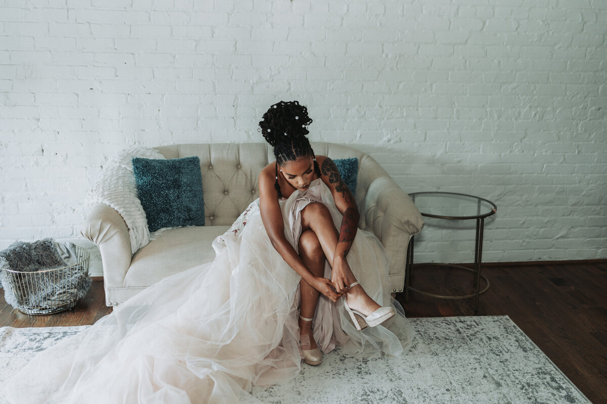 A bride is seen seated on a cream sofa, thoughtfully adjusting her high-heeled shoe, with her flowing gown draped around her, against a white brick wall backdrop at the Neon House in Norfolk, Virginia. Photographed as a stylish bridal editorial by Quinn Photography LLC.
