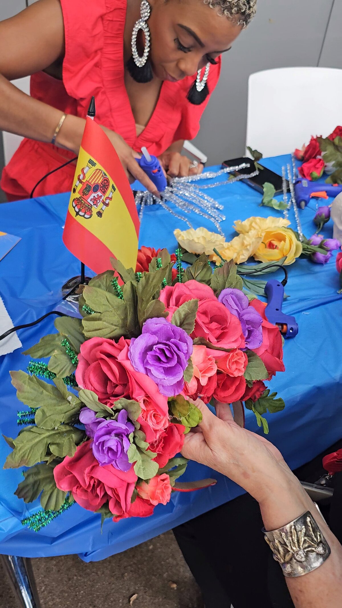 The world of craftsmanship as a woman in a stunning red dress delicately glues vibrant flowers onto a wreath in our artistic workshop. The long table is adorned with a spectrum of materials, and in the foreground, witness the beauty of the final piece—a masterpiece of colors and creativity.