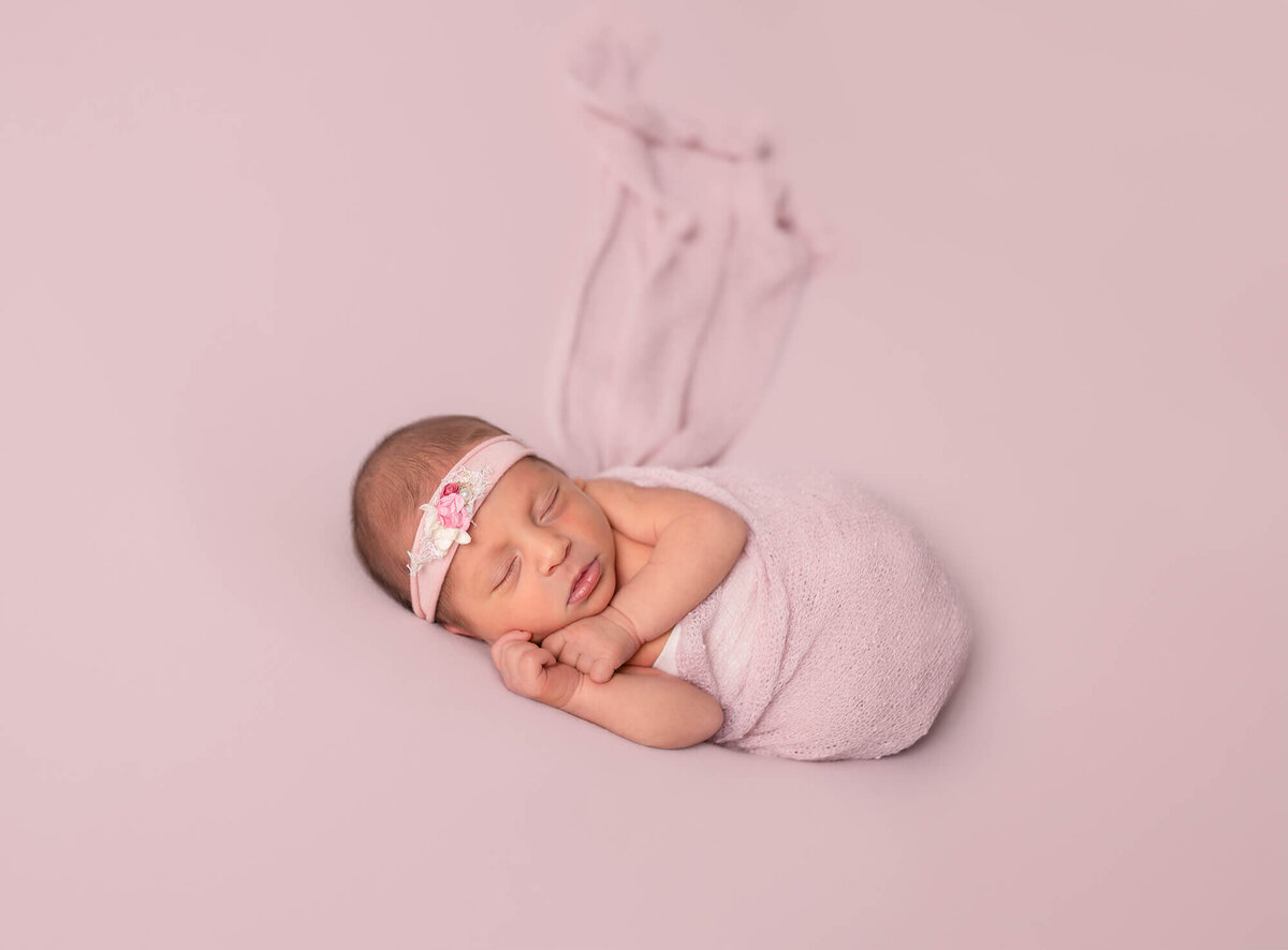 A newborn baby girl wrapped in a pink swaddle with a pink headband sleeps on a pink blanket