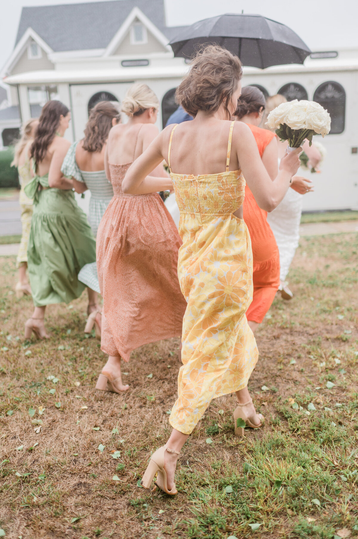 Bridesmaids in flowing dresses of green, orange, and yellow hurry across damp grass towards a vintage white trolley.
