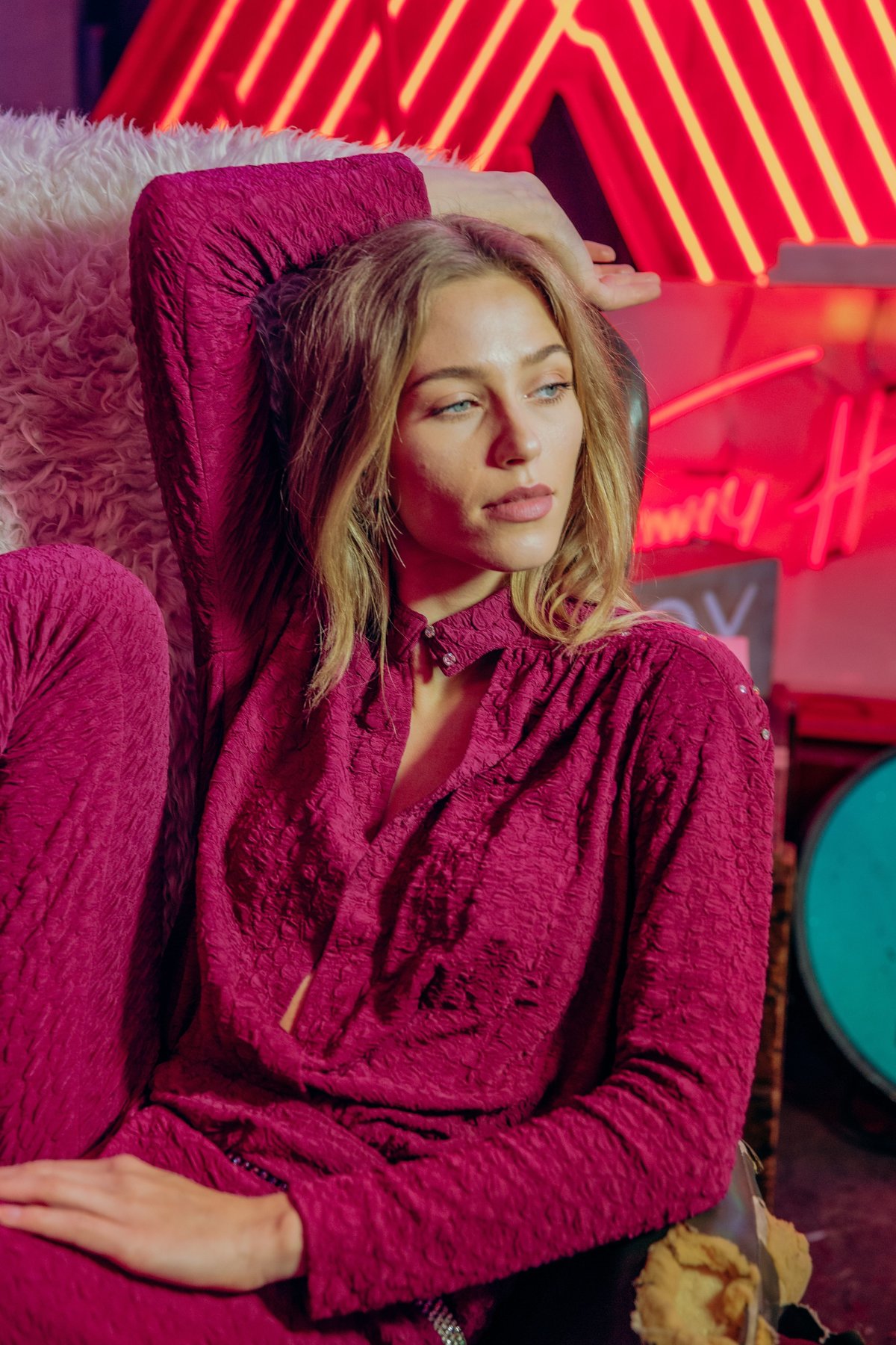 Anastazja wearing a pink jumpsuit, leaning back in a leather armchair, surrounded by neon signs