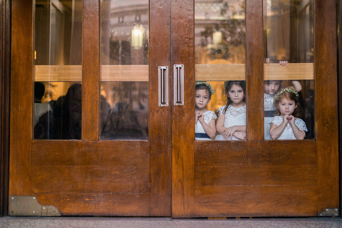Flower girls watching through a wooden door, anticipating the wedding ceremony, photographed by Rodrigo Varela Photography.