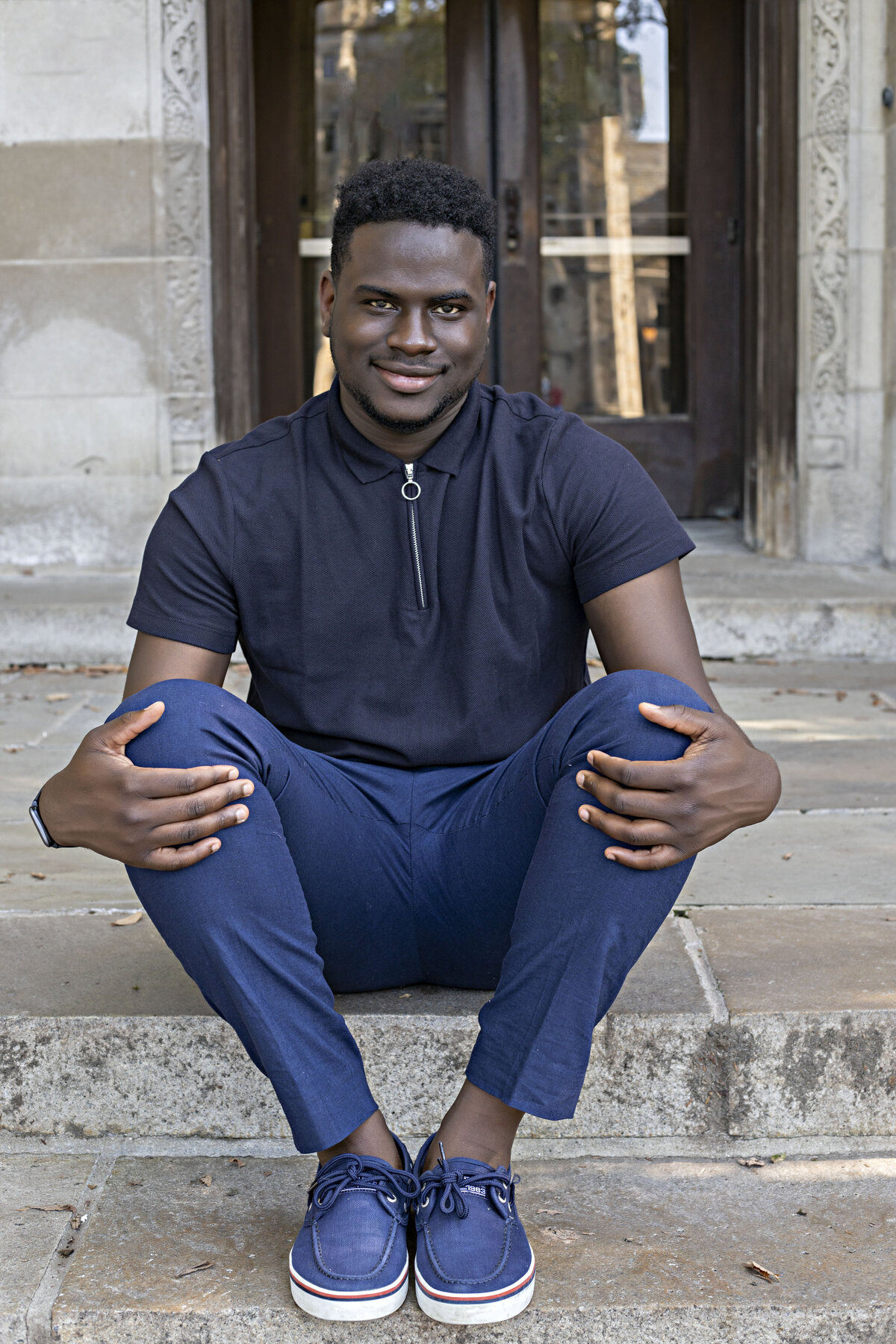 African American male wearing a navy blue zip up top, blue pants, and blue Sperry boat shoes sitting on steps in front of law library