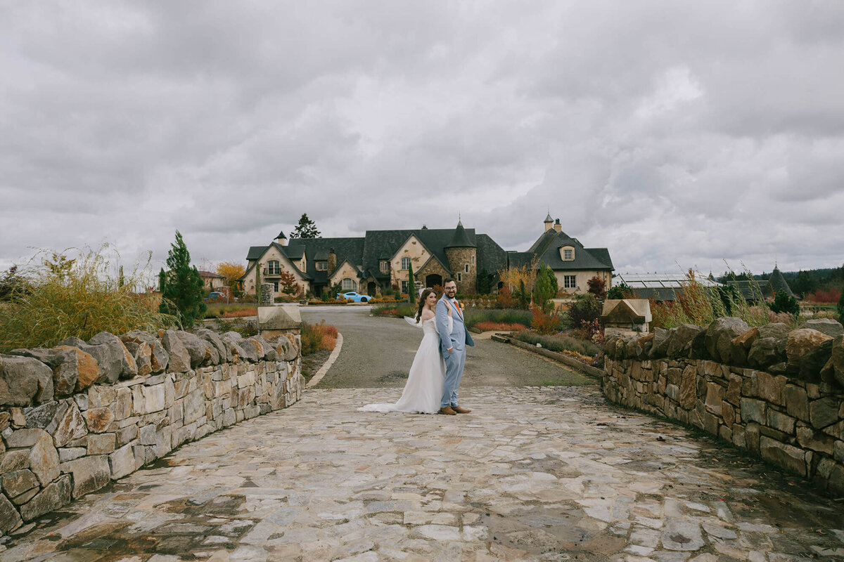 A bride and groom stand together in the driveway with the Chateau de Lis Wedding Venue in the Background.