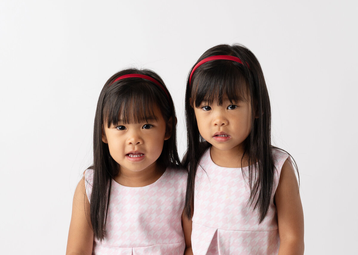 Twin girls with pink dresses and headbands posing for their school pictures.