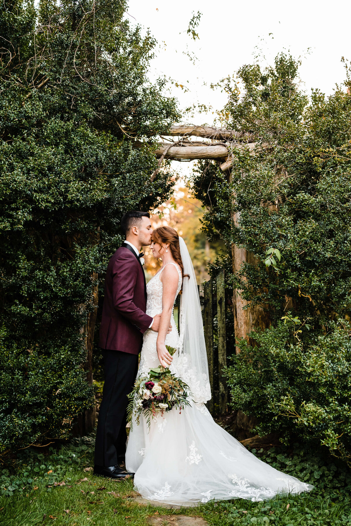 groom in a burgundy suit coat holds his bride by her waist and kisses her forehead as she leans into him and gently smiles in a secret garden at Murray Hill events for their Charlottesville wedding