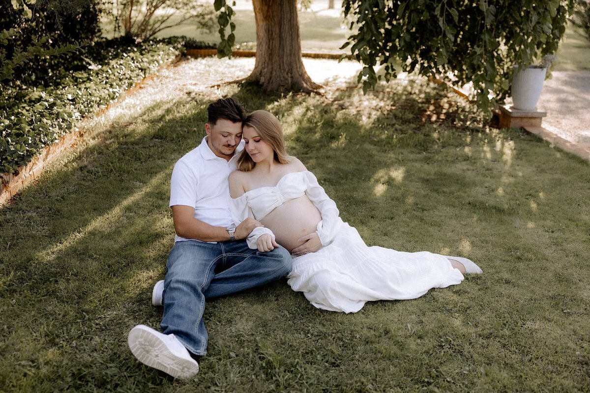 maternity couple sitting in grass area with eyes focused on her belly