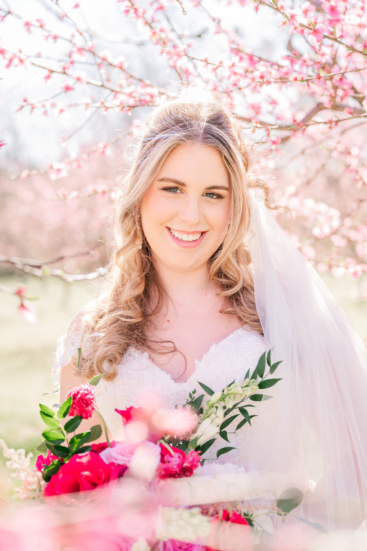 A glowing blonde bride holding a bright pink and red wedding bouquet surrounded by bright pink cherry blossoms enjoying her destination wedding photos 