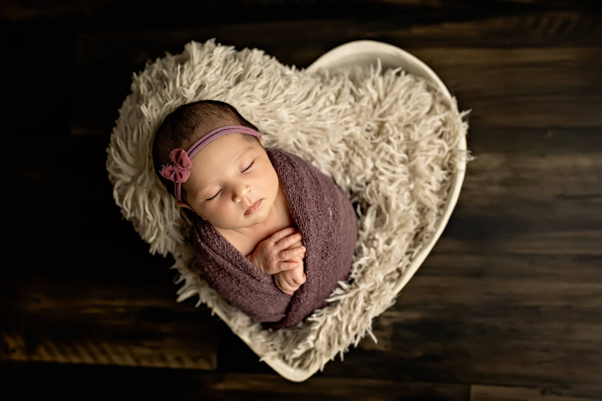A sleeping baby wrapped in a purple blanket, wearing a matching headband with a bow, is nestled in a heart-shaped basket lined with fluffy white fabric, placed on a dark wooden floor.