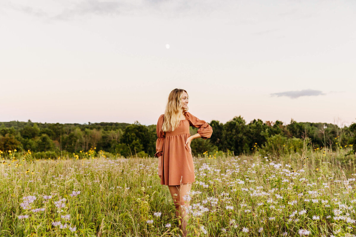 beautiful senior girl in an orange dress standing in a flower field near Green Bay as she looks over her shoulder