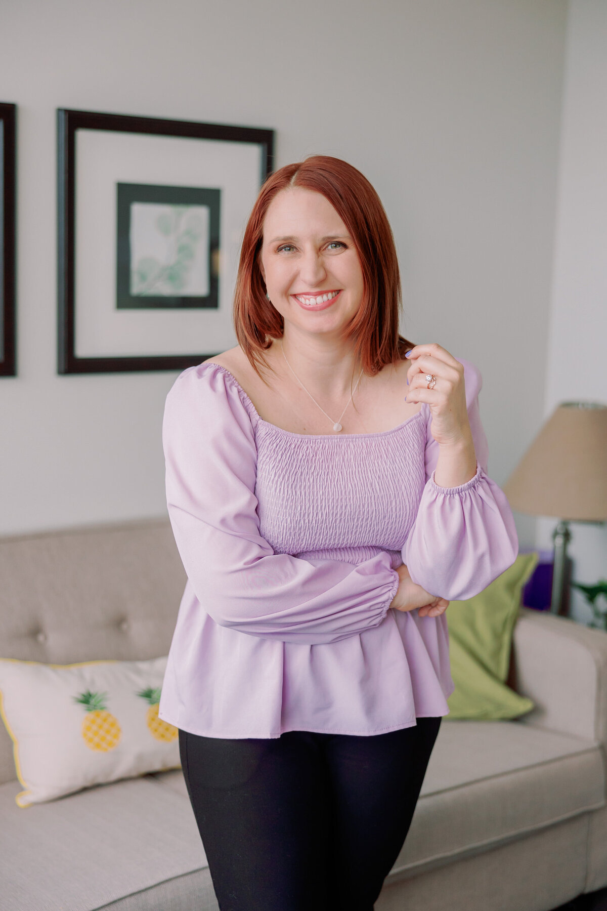 A person poses with their arms crossed indoors, a couch behind them.