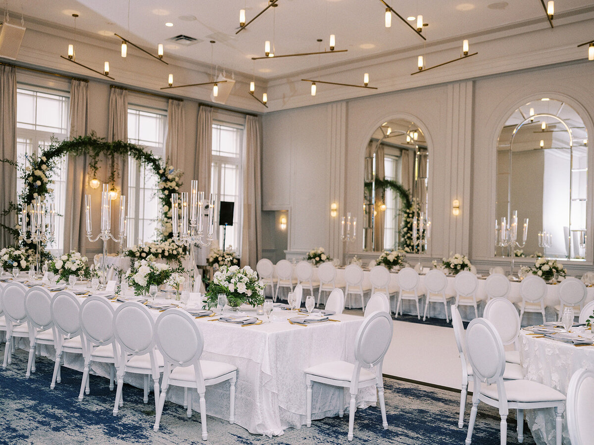 Elegant banquet hall adorned with a floral arch, white tablecloths, white chairs, large windows, and modern light fixtures. Tables are set with glassware and floral arrangements, creating the perfect setting for a cultural Calgary wedding at Fairmont Palliser.