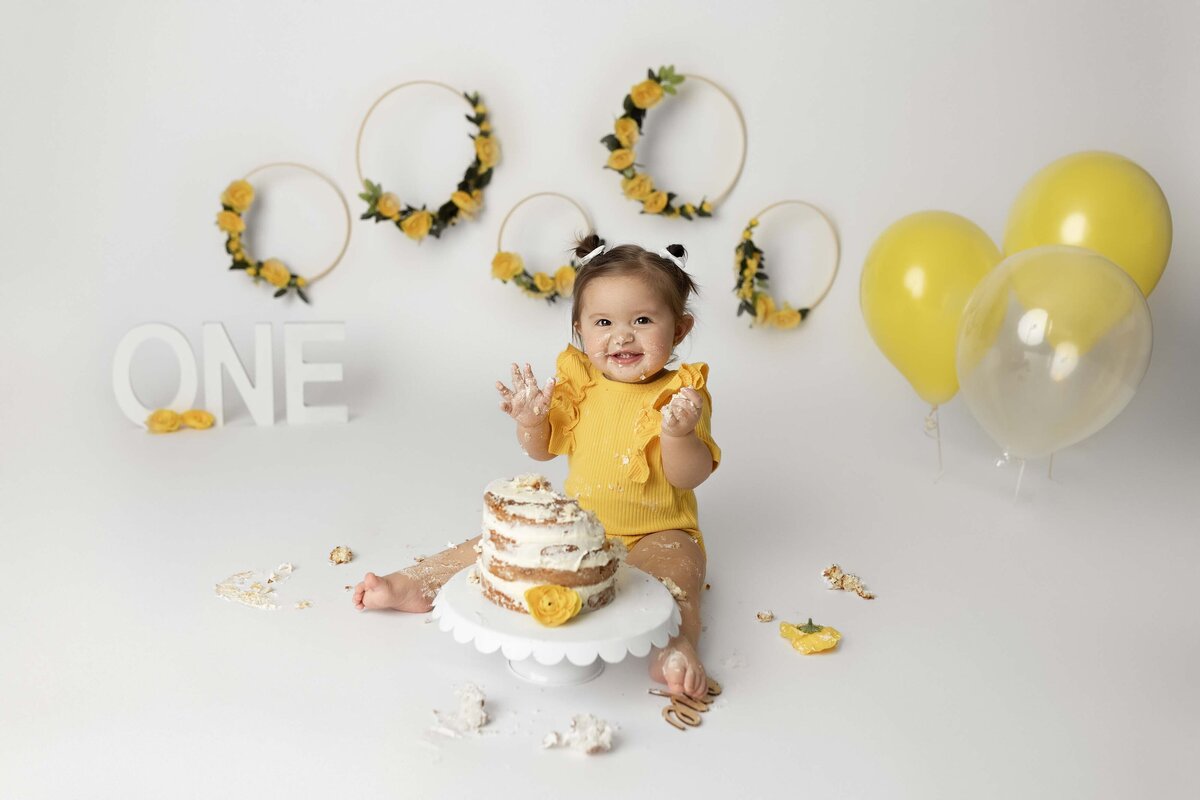 Cake smash first birthday photos by top London, Ontario baby photographer Amy Perrin-Ogg. Baby girl in a yellow romper has cake on her hands and smiling at the camera. There are lemons on the cake with yellow balloons and yellow floral hoops in the background.