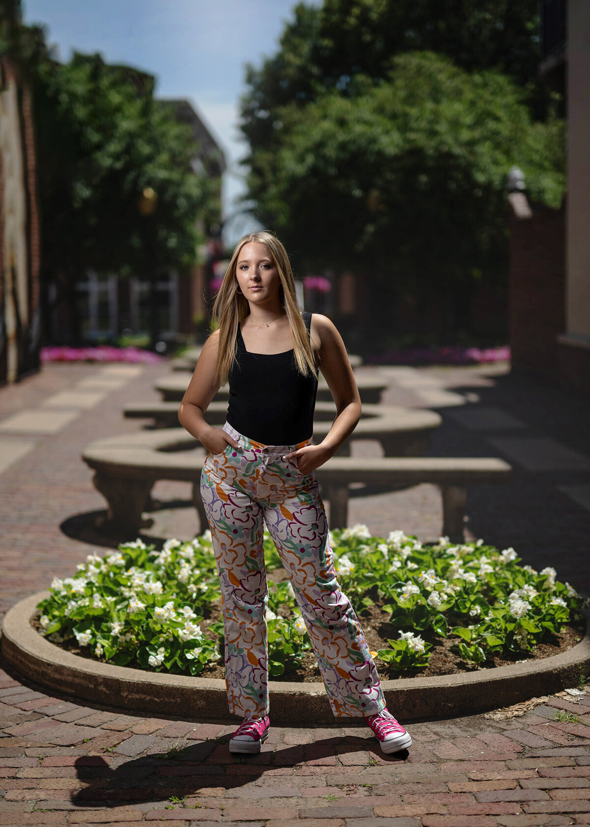 A high school senior stands in a park path by a flower bed with hands in her colorful pants pockets