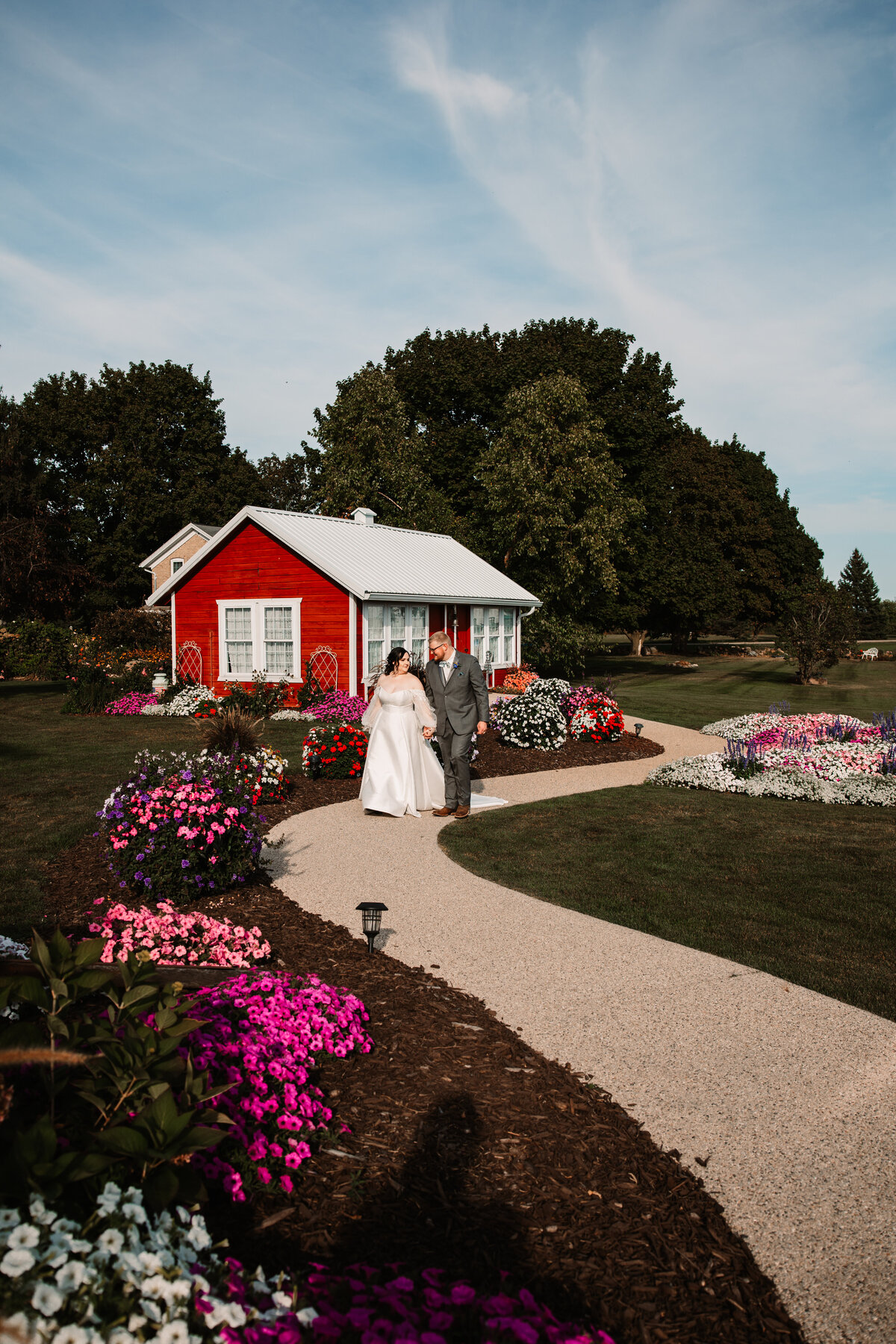 Newlyweds walk together on a garden sidewalk lined with flowers away from a small red cottage