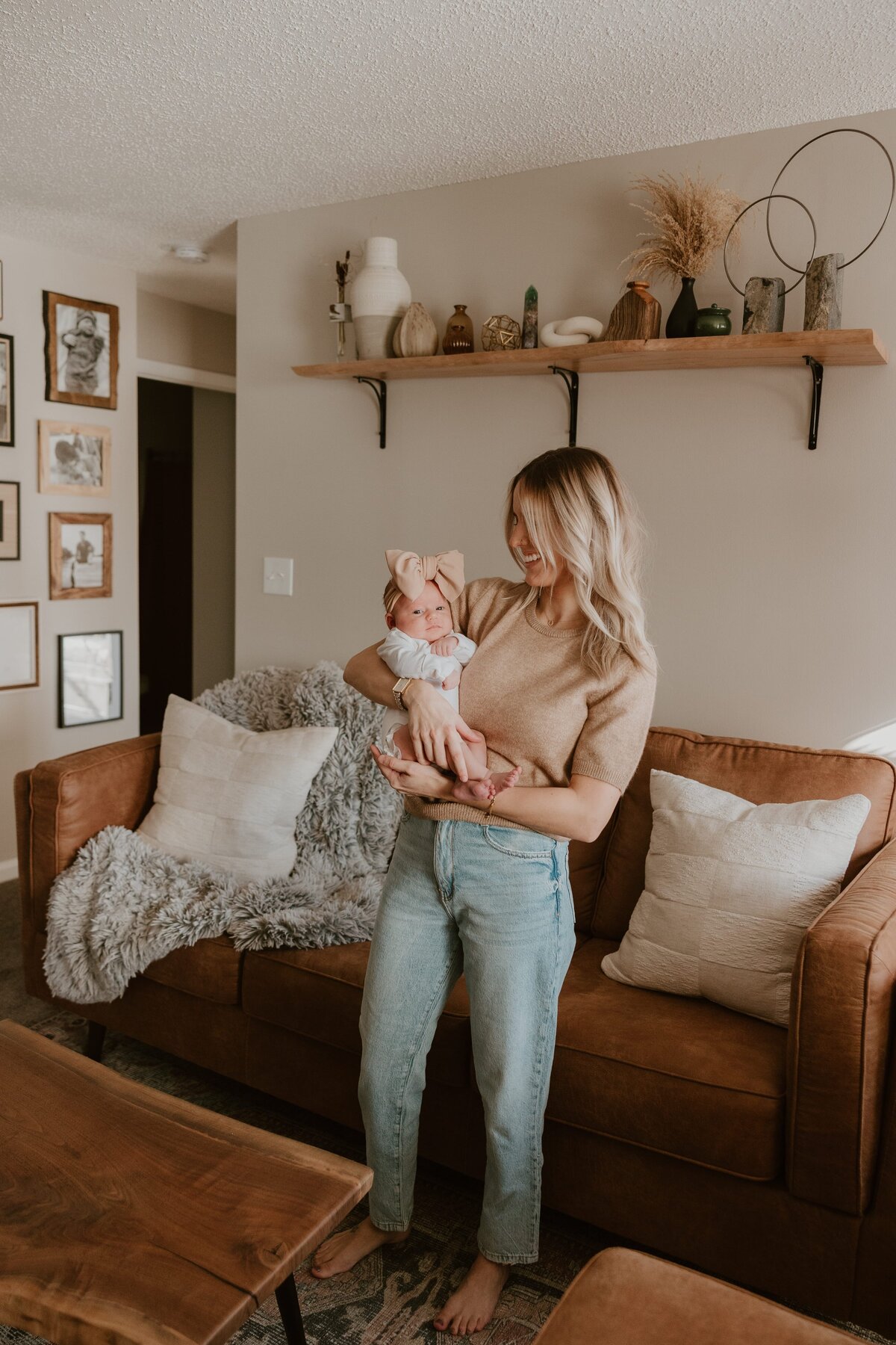 A mother with long blonde hair, wearing a beige short-sleeve sweater and light-wash jeans, holds her baby girl in a cozy living room. The baby is dressed in white with a large beige bow headband. The mother smiles while looking down at her baby. The living room features a brown leather couch with neutral pillows and a gray textured blanket, a wooden coffee table, a floating wooden shelf with decorative items, and a gallery wall of framed family photos.