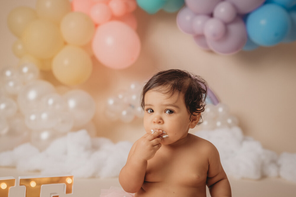 Photo shoot at Atlanta, Georgia cake smash photographer studio with one year old little girl in pink tutu smashing a cake with rainbow pastel balloon garland behind her with white fluffy clouds.