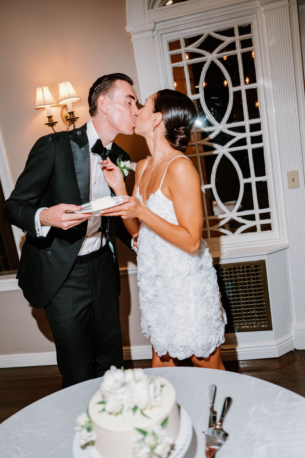 Rhode Island Wedding Photographers |A couple in formal attire shares a kiss at a wedding reception. The groom holds a plate, and the bride wears a white dress, smiling in a warmly lit room.