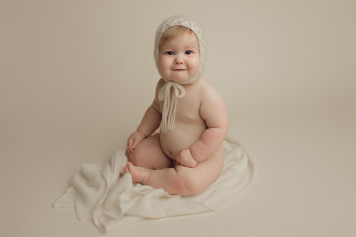 one year old girl wearing a white bonnet sitting and looking at the camera