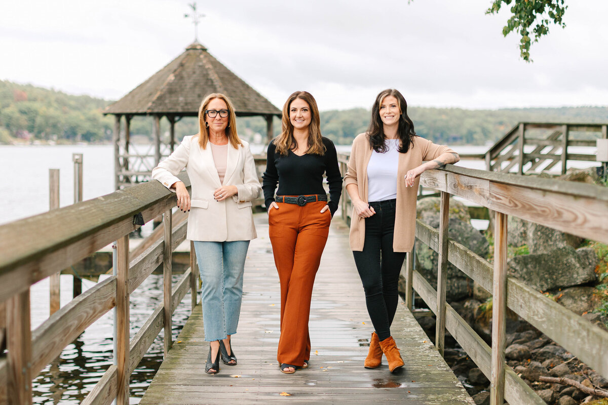 Three professional women stand confidently on a wooden dock with a gazebo, overlooking a calm lake. They are dressed in stylish neutral and earth-toned outfits, exuding a poised and approachable presence against the scenic waterfront backdrop.