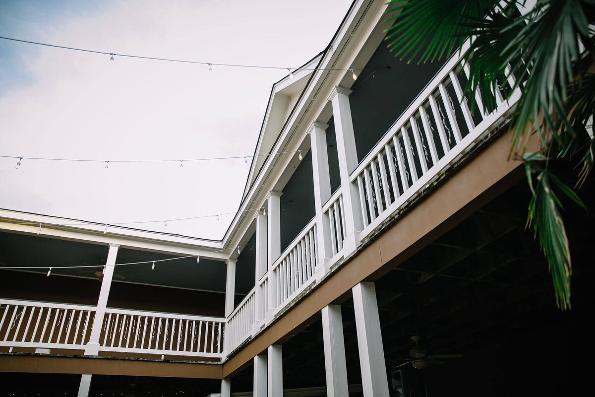 A low-angle view of a building with white railings, columns, and balconies, featuring string lights overhead and a palm leaf partially visible on the right side. The sky is clear.
