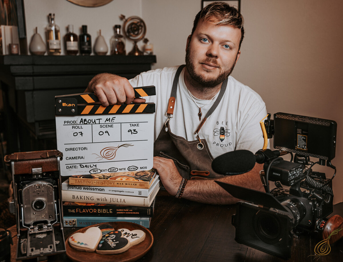 A man sits at a table, holding a film clapperboard that reads "About Me." He wears a white shirt with a bee logo and "Bee Proud" text, along with a grey apron. Surrounding him are a vintage camera, modern video equipment, and a stack of food-related books, including titles like *Tasting History* and *Baking with Julia*. A plate of decorated cookies sits in the foreground. The scene is cozy, with bottles and decor in the background, suggesting a storytelling and culinary focus.