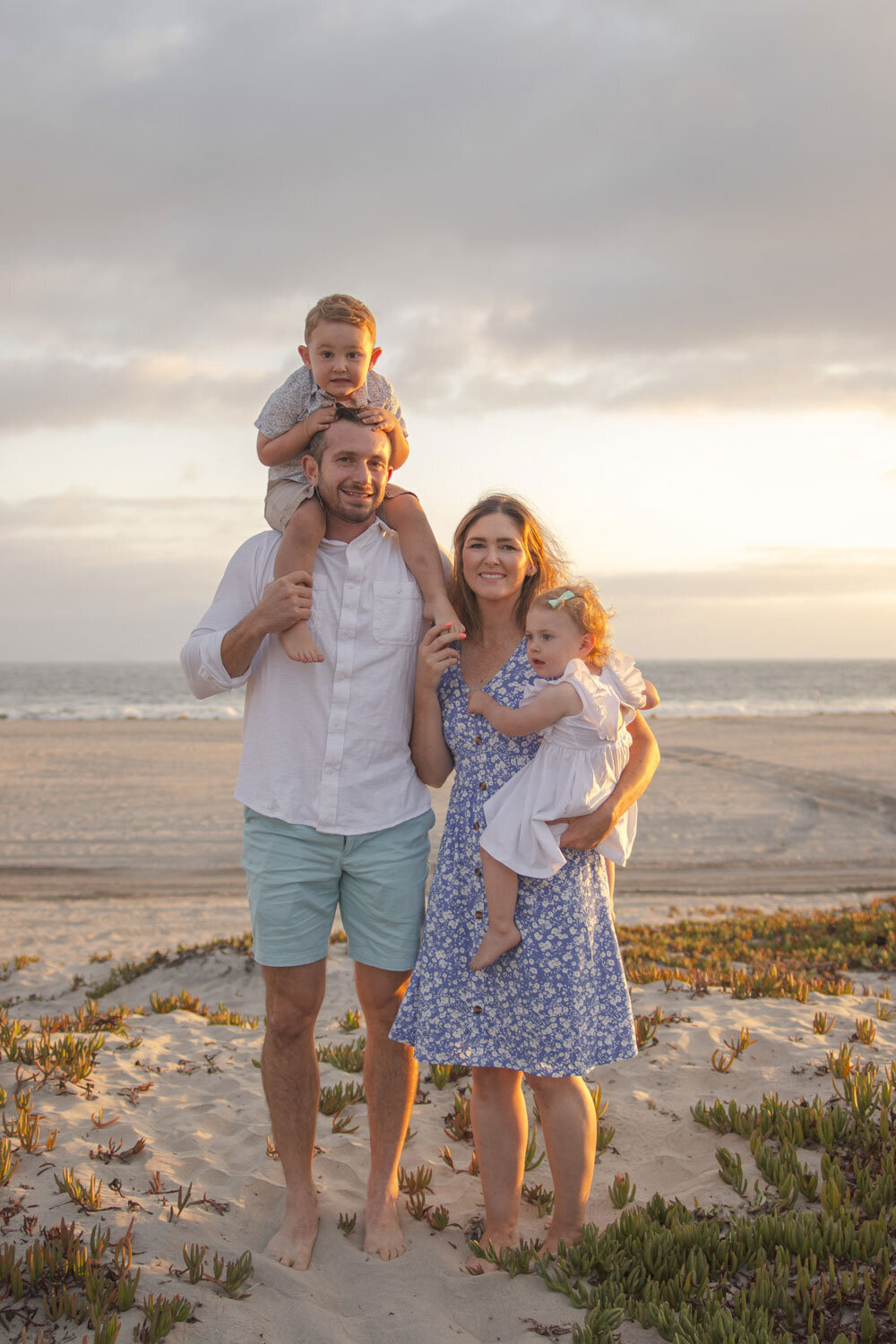 A happy family standing on a beach, with a father carrying his young son on his shoulders and the mother holding their daughter in her arms at sunset