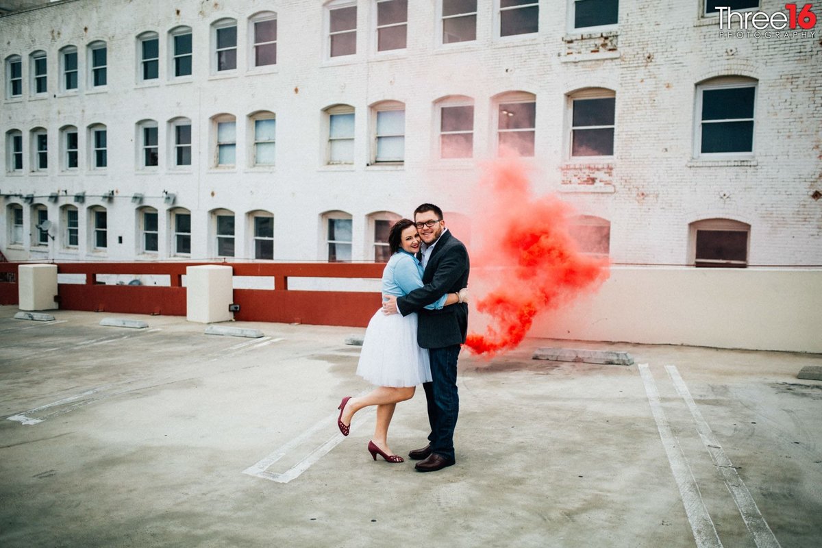 Red paint bomb goes off behind engaged couple as they pose for photos on the top of a parking structure