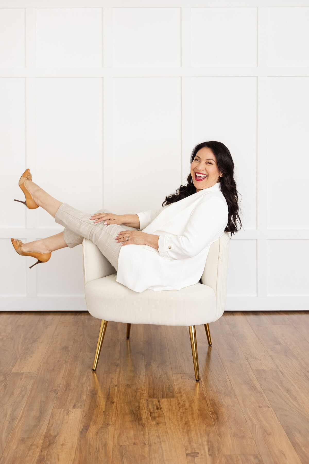 Woman wearing white blazer and tan pants with brown heels sitting on a white art deco chair with her legs kicked up for her brand photoshoot