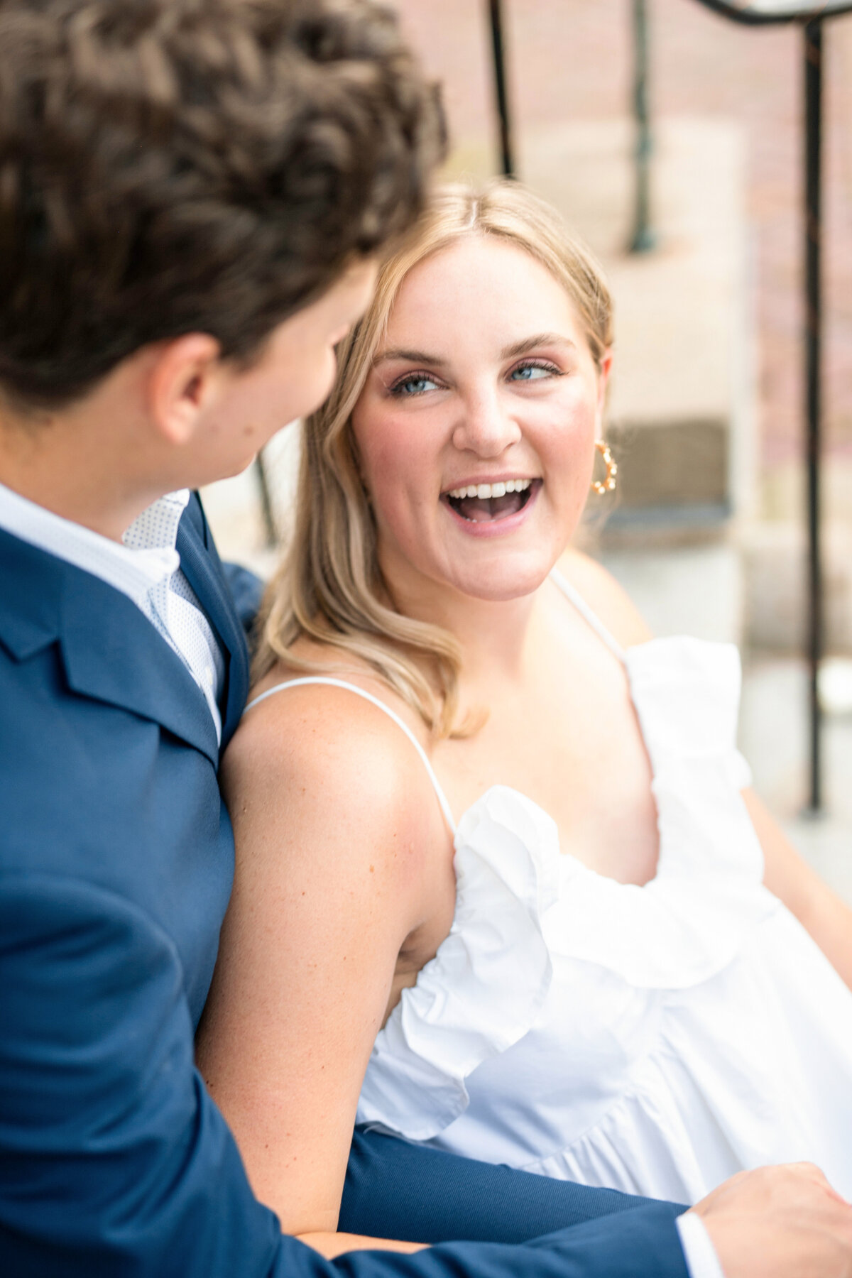 A couple sits together outdoors, smiling and looking at each other. The woman, wearing a white dress, has blonde hair and is laughing. The man, wearing a blue suit, gazes at her affectionately. They appear to be sitting on steps.