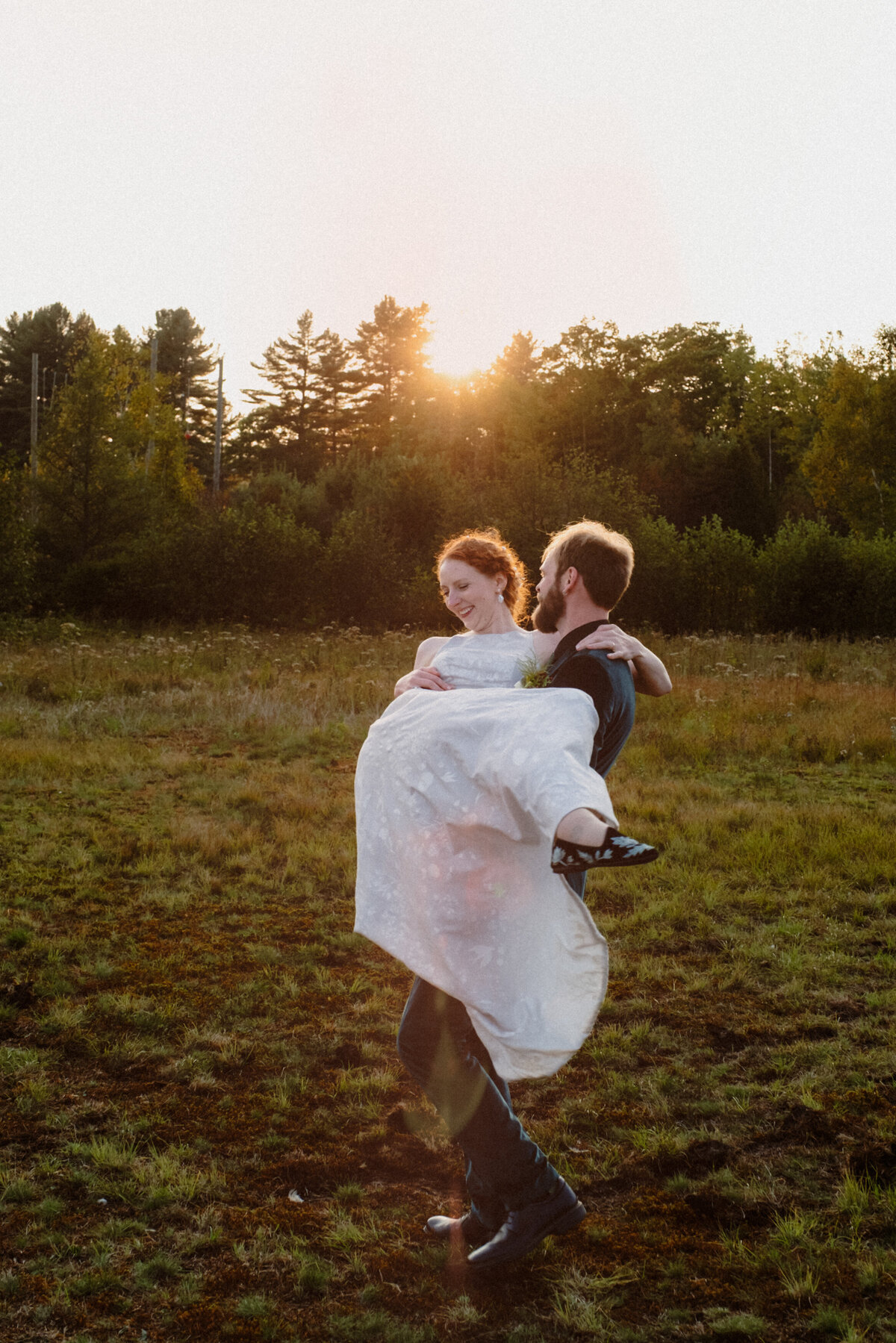 groom holding bride in his arms in a field at sunset