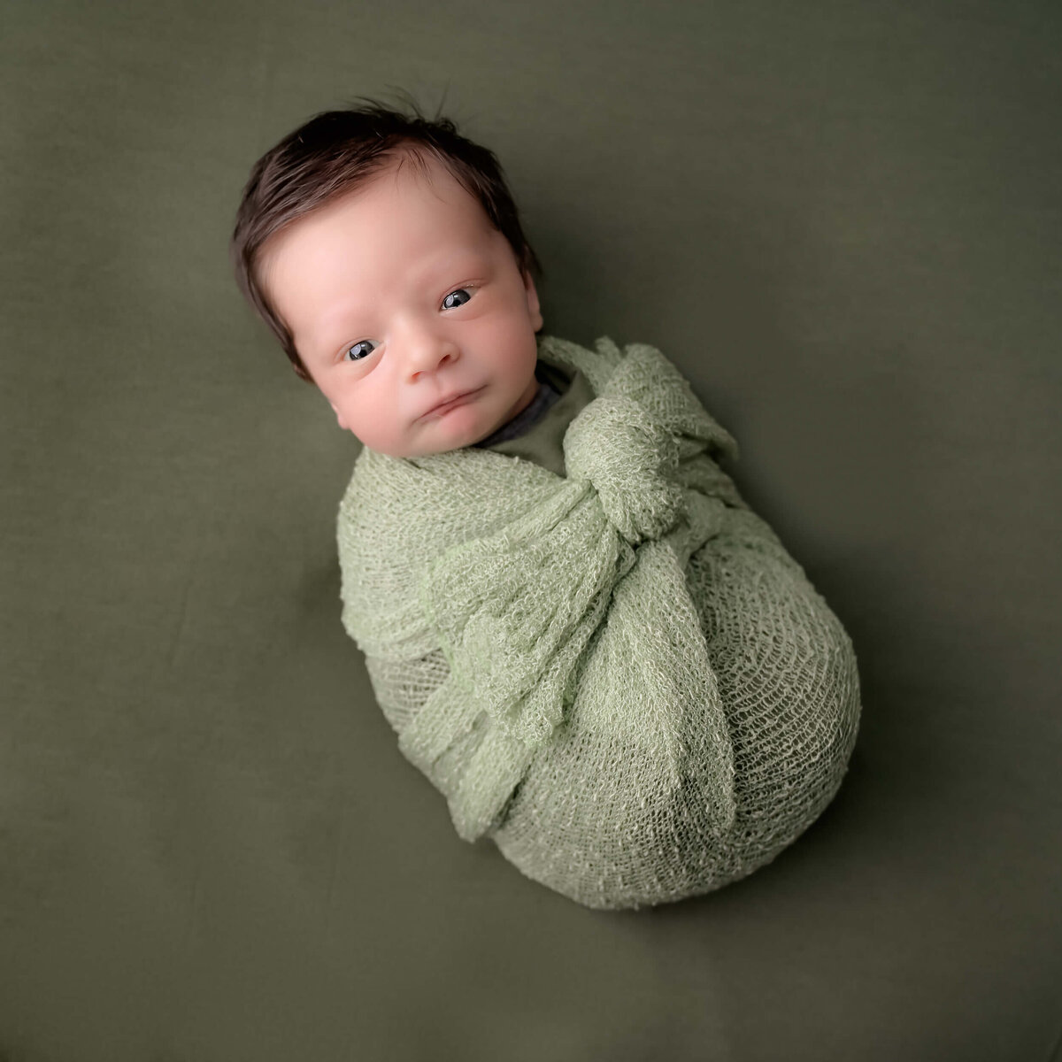 Newborn photo session by Beth Benecki of newborn with eyes wide open and he is wrapped in green  and laying on a green background