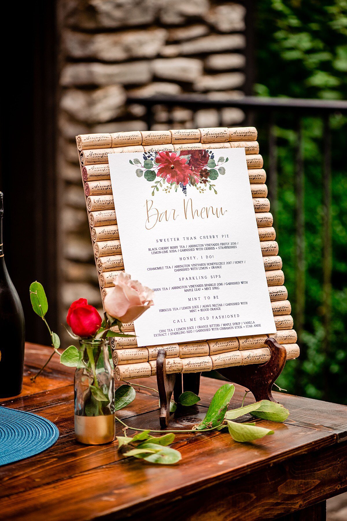 Signature cocktail bar menu decorated with red and burgundy flowers and greenery attached to a background made of wine corks on a small easel. The bar sign sits on a wood farm table with a glass and gold bud vase of red and blush roses at Arrington Vineyards.