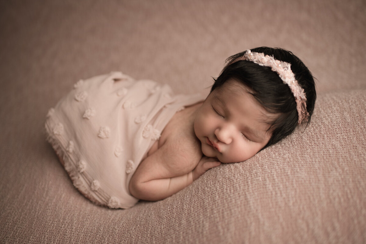 Side profile image of Riverside, CA newborn photoshoot. Baby girl sleeping on her belly and wearing a pale blush headband. Pale blush fabric is draped across her back. Her hands are folded under her cheek.  Captured by Best Riverside, CA newborn photographer Bonny Lynn Photography.