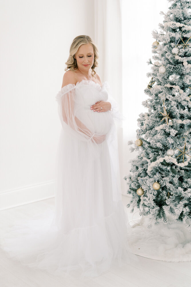 expecting-mom-in-white-gown-next-to-flocked-christmas-tree