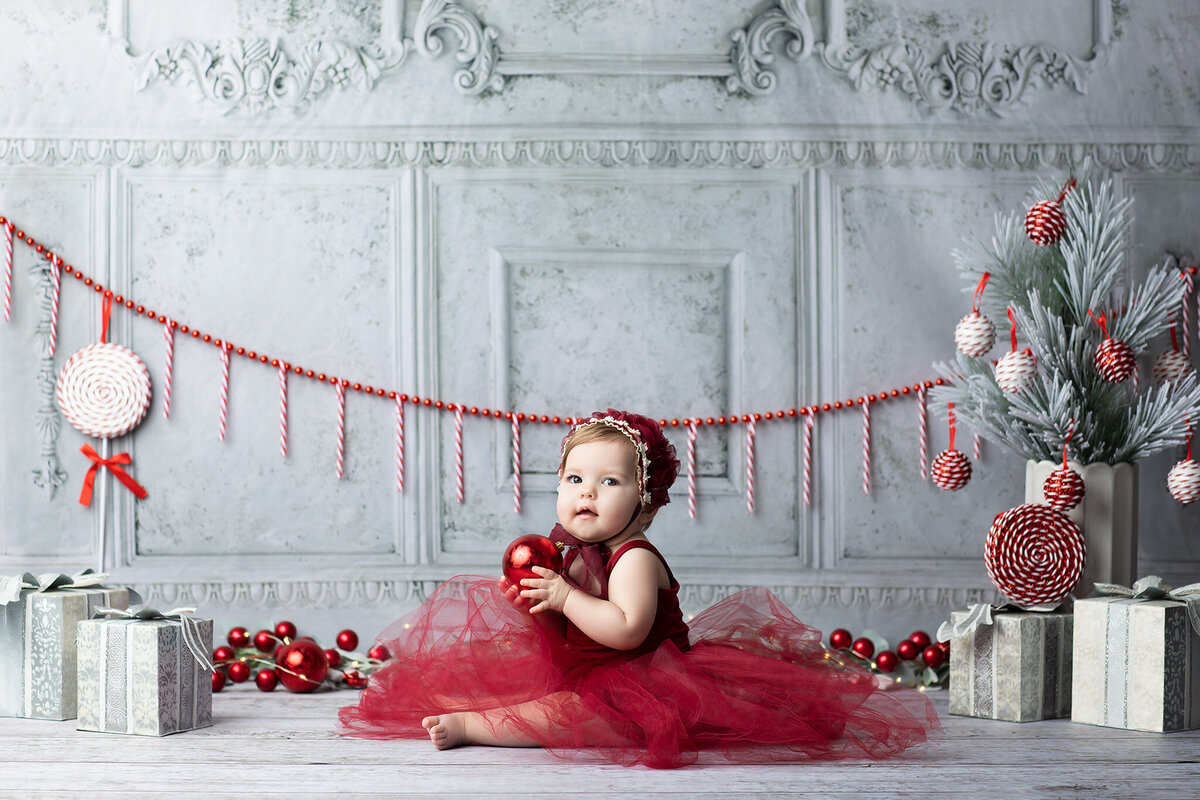 Baby girl in a deep red tulle dress holding a Christmas ornament, surrounded by festive candy-themed décor, photographed by a Dallas milestone photographer.
