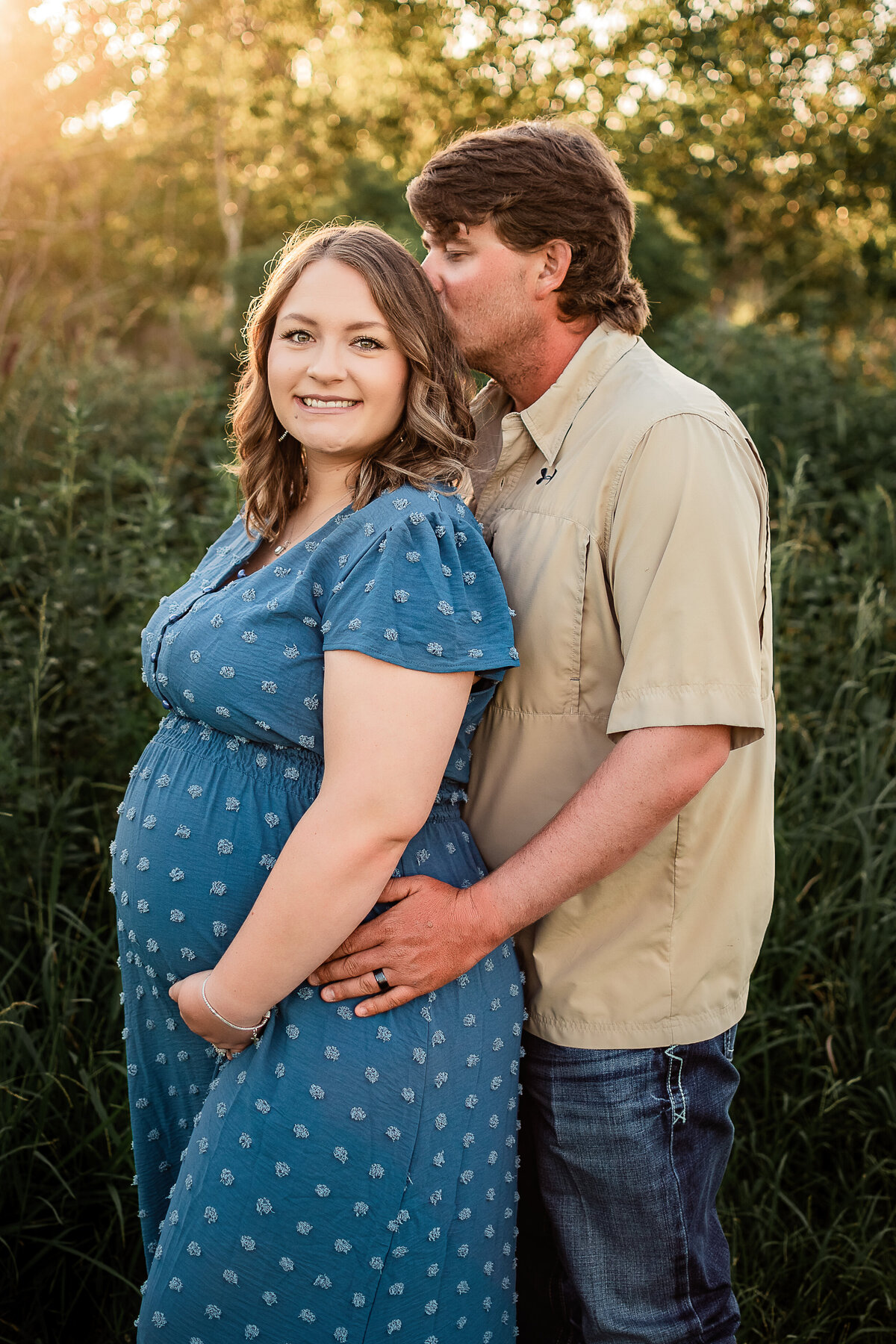 A husband stands behind his wife and kisses her head as she holds her baby bump and smiles.