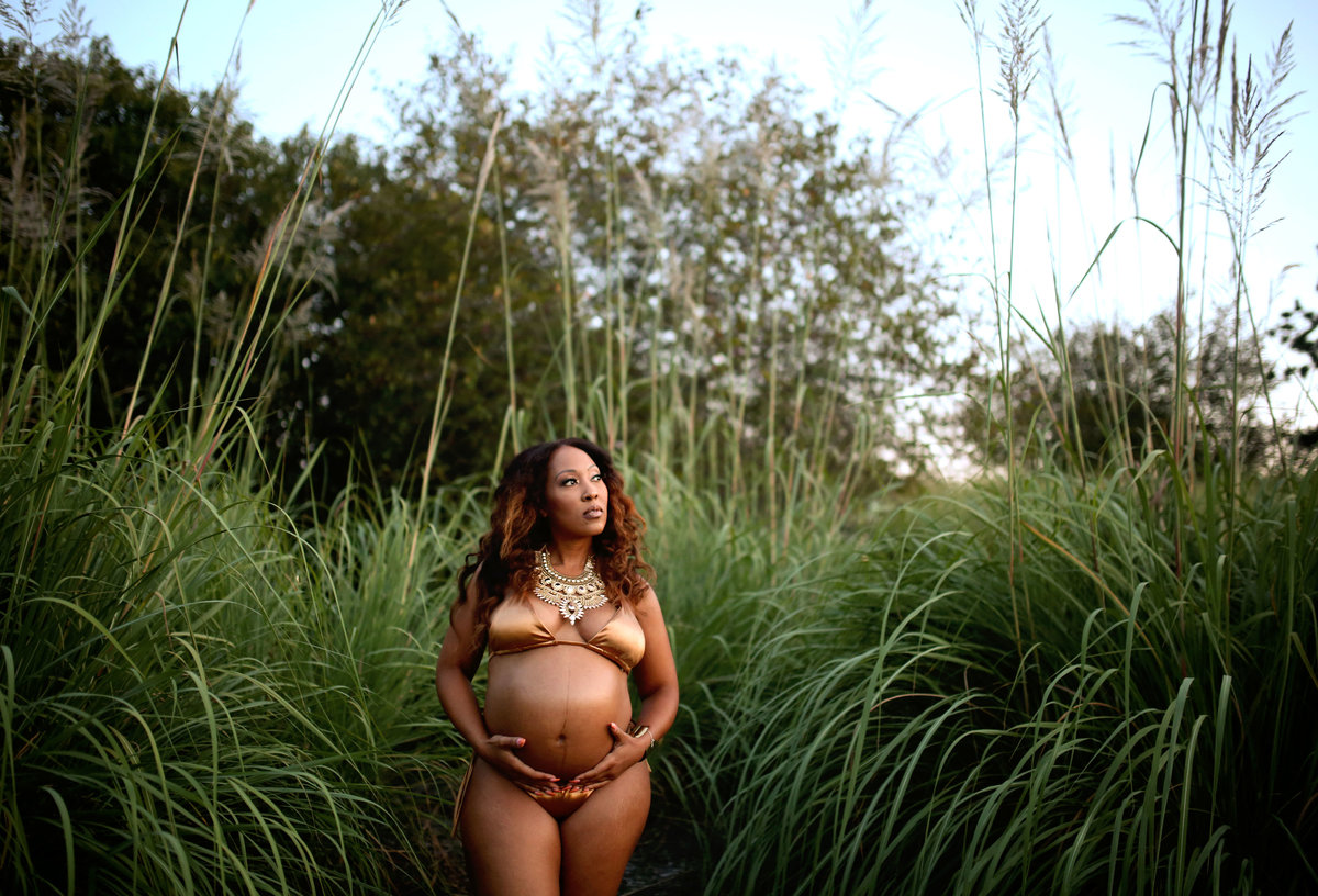 black pregnant woman in metallic gold bathing suit outdoors surrounded by lush green foliage