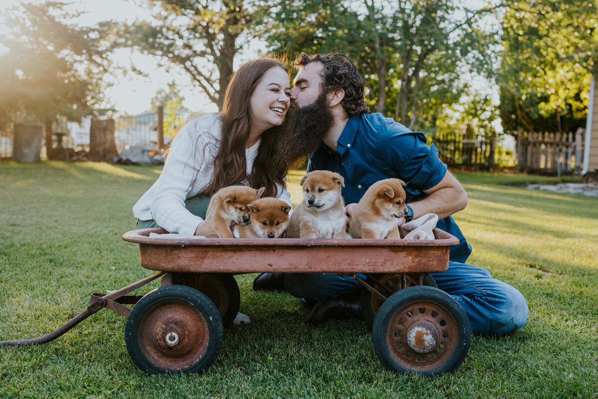 Couple leans in for kiss while four puppies sit in wagon in front.