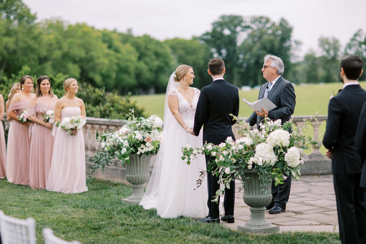 View of a wedding ceremony at the Great Marsh Estate with a bride and groom holding hands while a priest officiates the ceremony