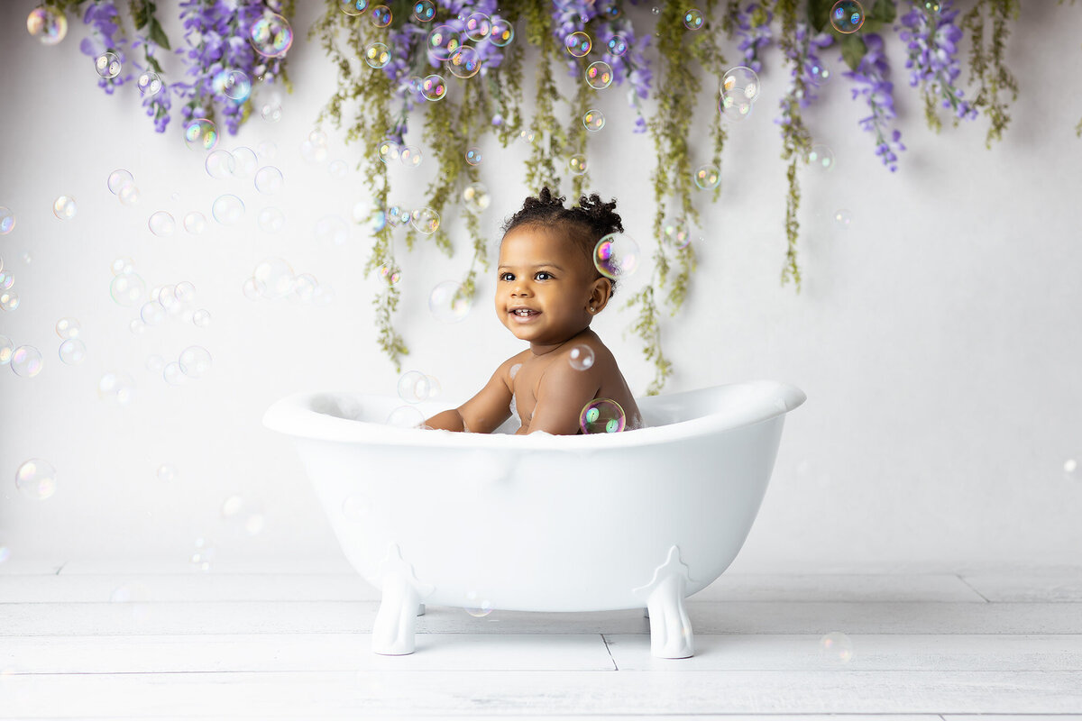 A smiling toddler enjoys a bubble bath surrounded by floating bubbles and cascading lavender florals during a DFW milestone photography session.