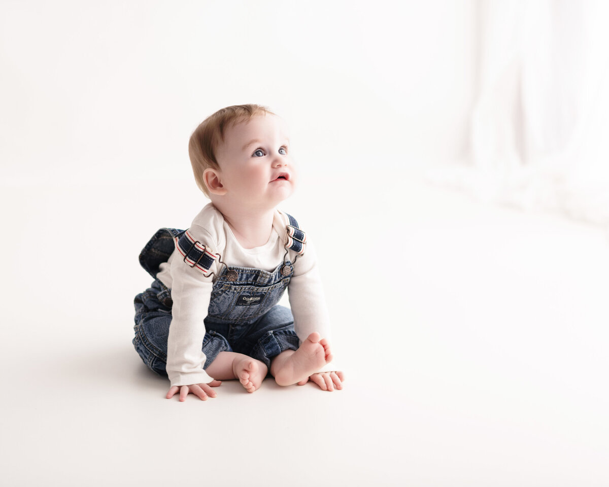 Clean milestone portrait of a baby in overalls with a white background—capturing childhood curiosity and innocence.