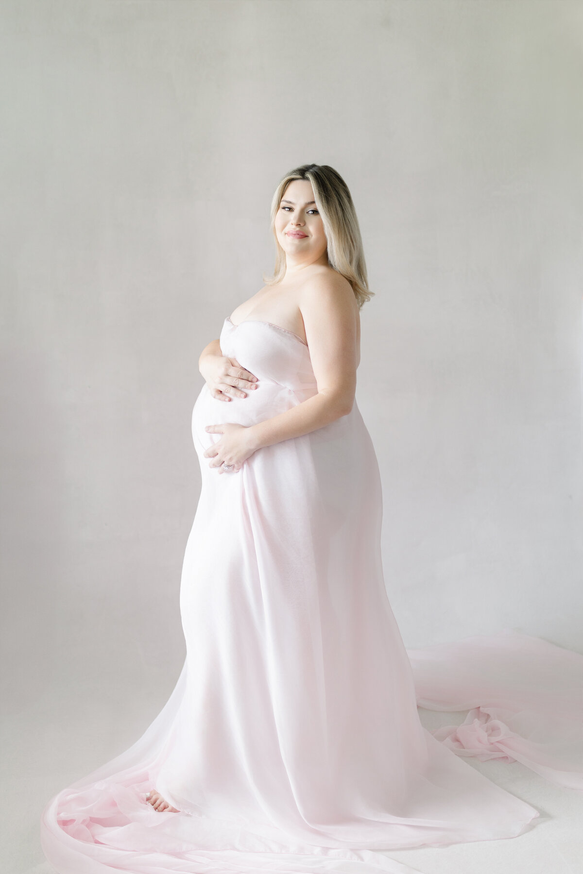 Elegant maternity image of Calli holding baby bump in minimalist white studio wearing blush pink