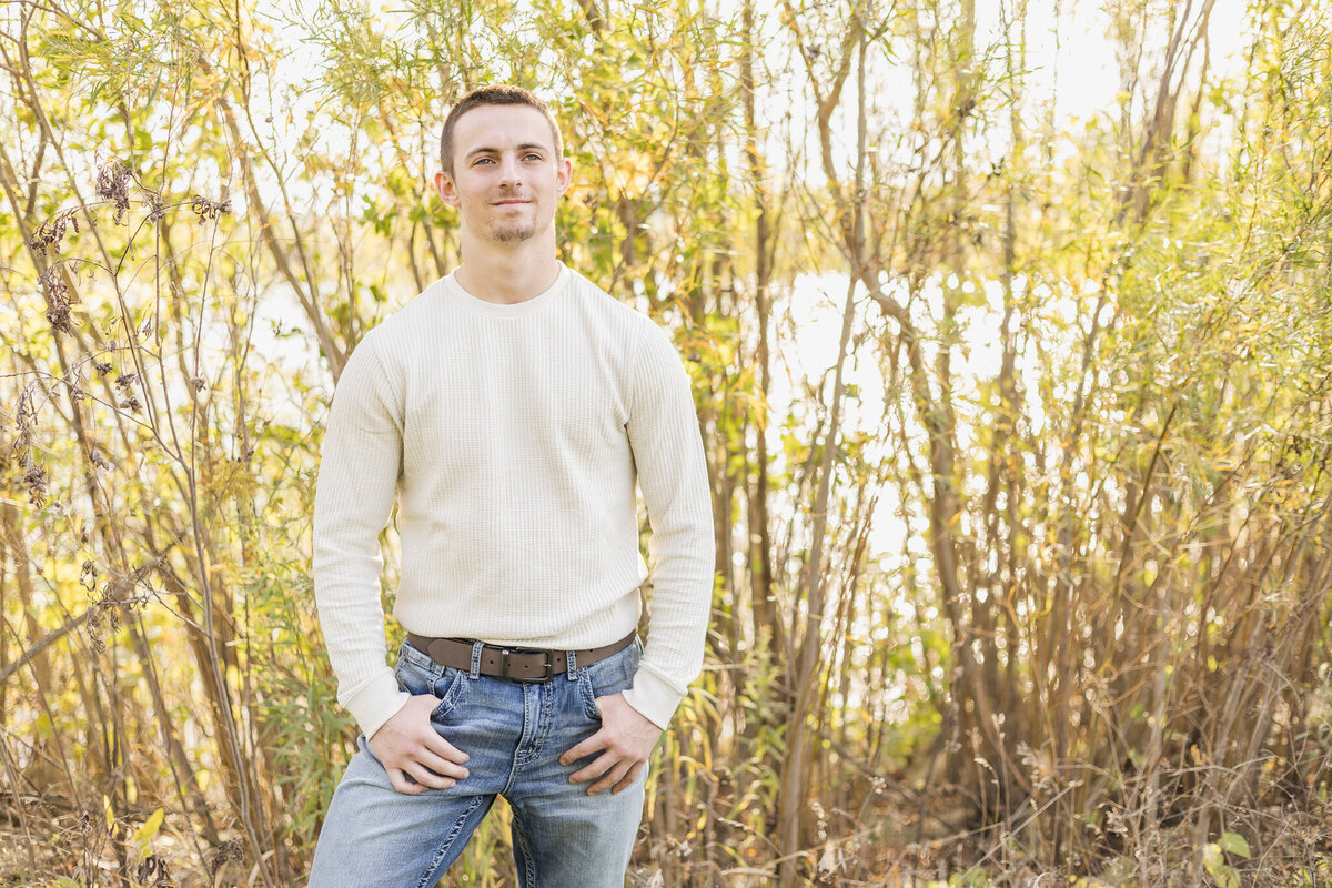 high school senior male, standing in grass, with trees in the background, thumbs in his pockets, wearing a light tan long sleeve shirt, blue jeans, a belt, and looking away from the camera with a soft smile