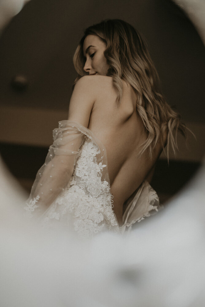 image shot through a round makeup mirror, of a blond girl in a wedding dress that is unbuttoned and falling halfway down her torso, expoing her back. She is looking down over her shoulder.