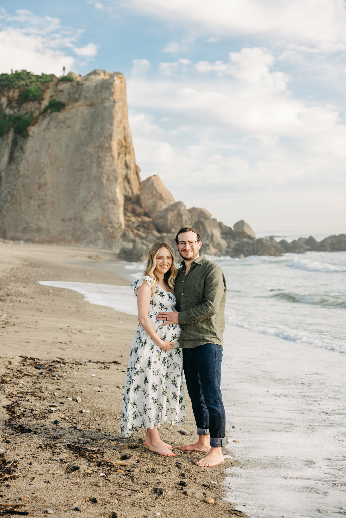 Pregnant woman and husband cradle pregnant belly while smiling at the camera on the beach in Malibu.