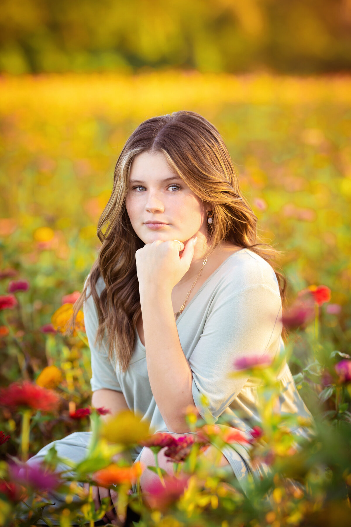 Senior girl with long brown hair crouched down in a zinnia field.  The flowers are pink, orange, and yellow.  There is lots of sunlight behind her.  She is looking at the camera with one hand under her chin.