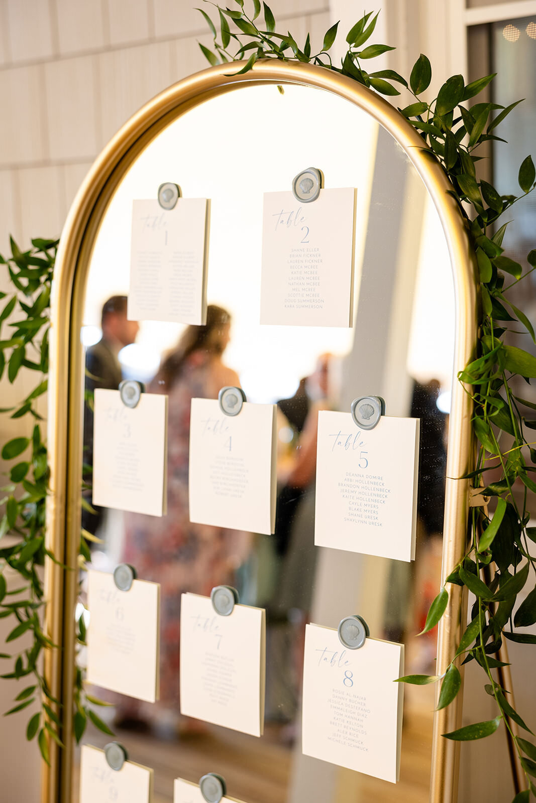 A seating arrangement display on a gold-framed mirror adorned with greenery, with wedding guests reflected in the background.
